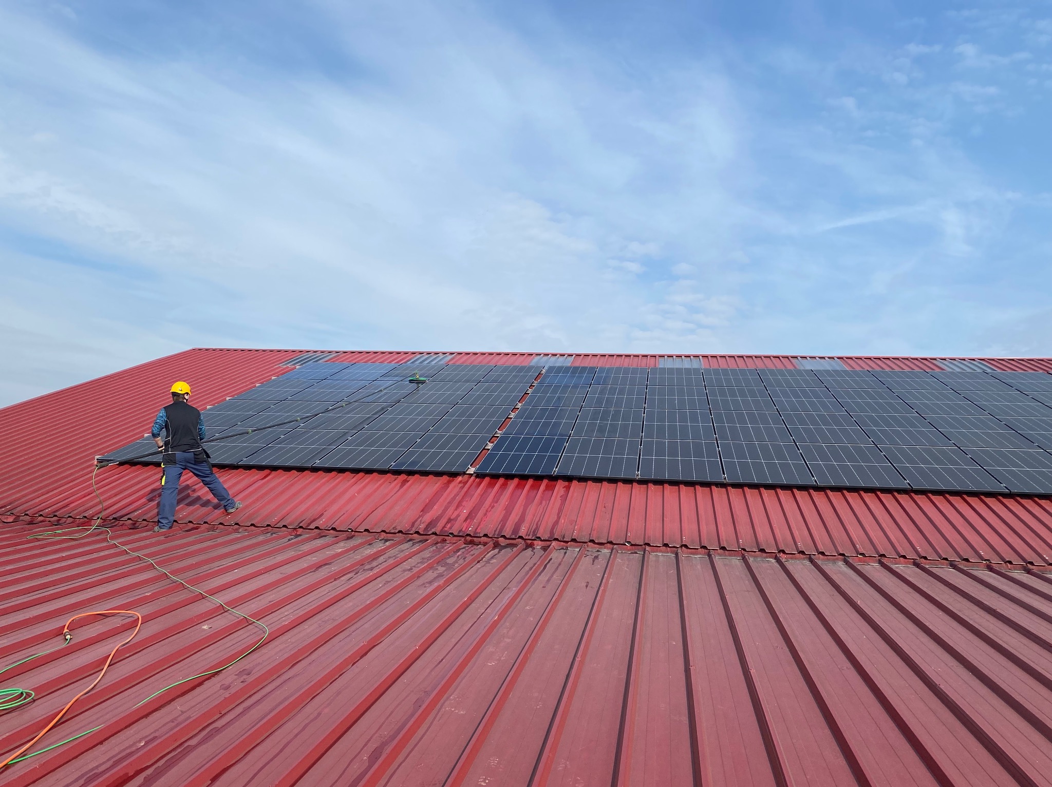 Pracownik w kasku czyści panele słoneczne na czerwonym, metalowym dachu za pomocą długiej szczotki. Widoczne węże doprowadzające wodę i błękitne niebo.