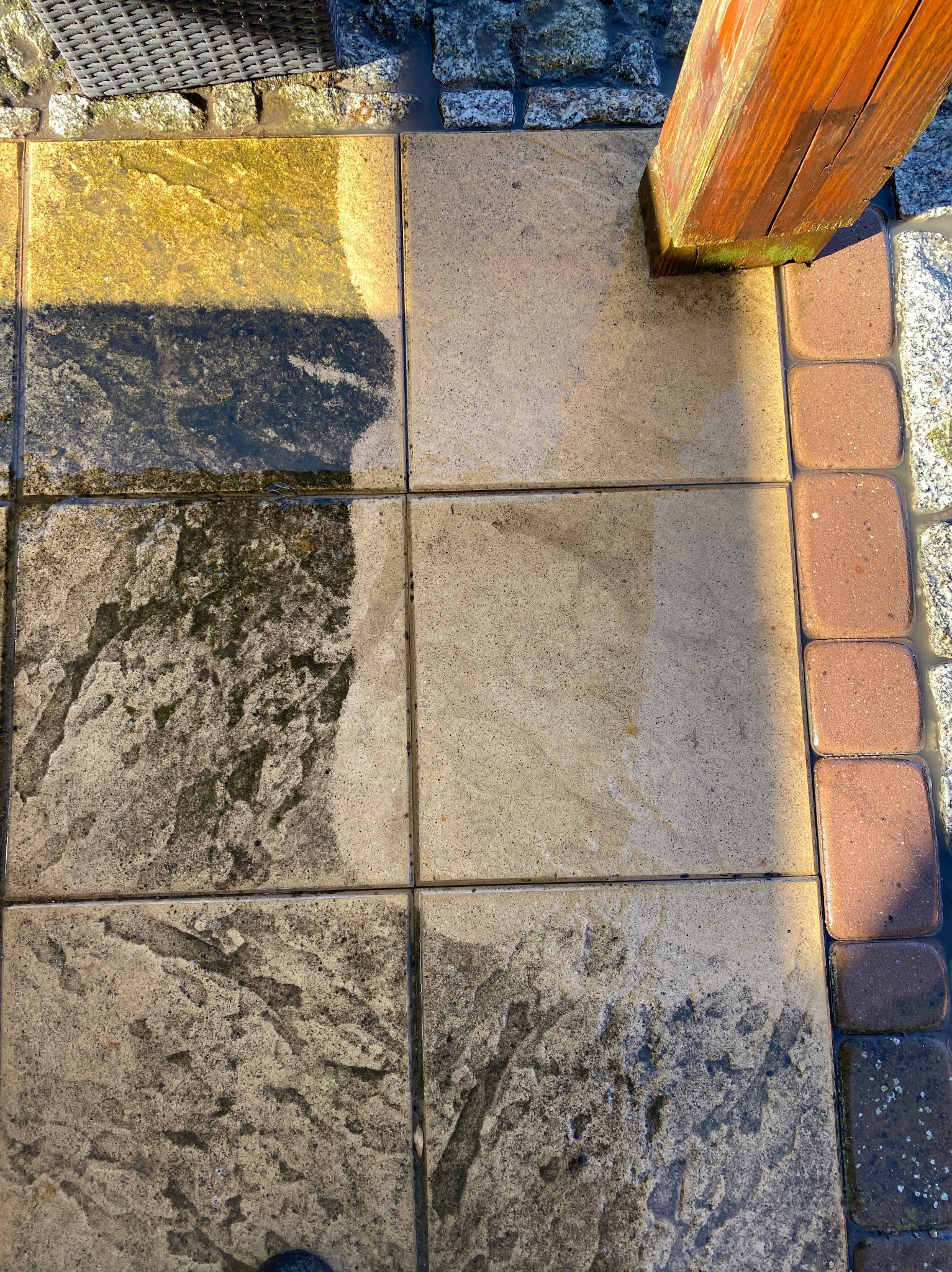 Częściowo umyta kostka brukowa w słoneczny dzień, widoczna różnica między czystą a zabrudzoną powierzchnią. Obok drewniany słupek i obrzeże z kostki.