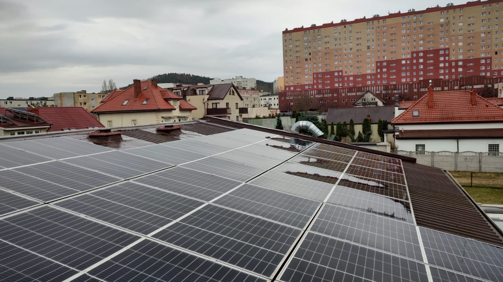 Błyszczące panele słoneczne na brązowym dachu budynku w pochmurny dzień. W tle zabudowa miejska i wzgórze.