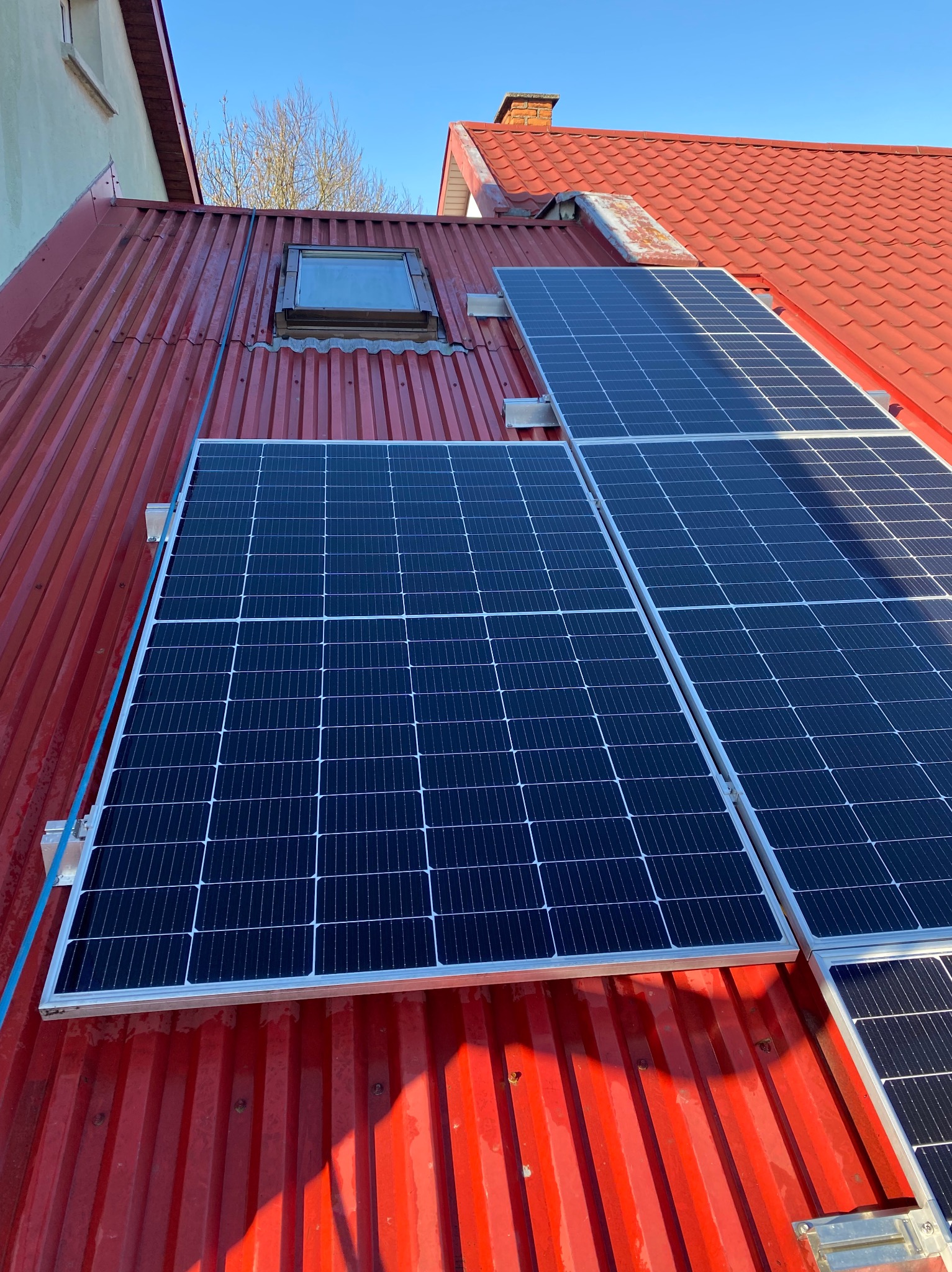 Czerwony dach z panelami słonecznymi w słoneczny dzień. Widoczny fragment okna dachowego i komin. Ujęcie z dołu, podkreślające instalację paneli.