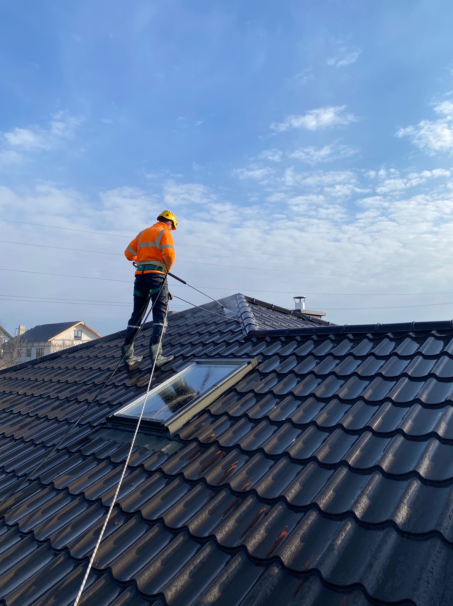 Pracownik w kasku i kombinezonie czyści dachówkę z okien połaciowych za pomocą myjki ciśnieniowej. Widok z dołu na błękitne niebo z chmurami.