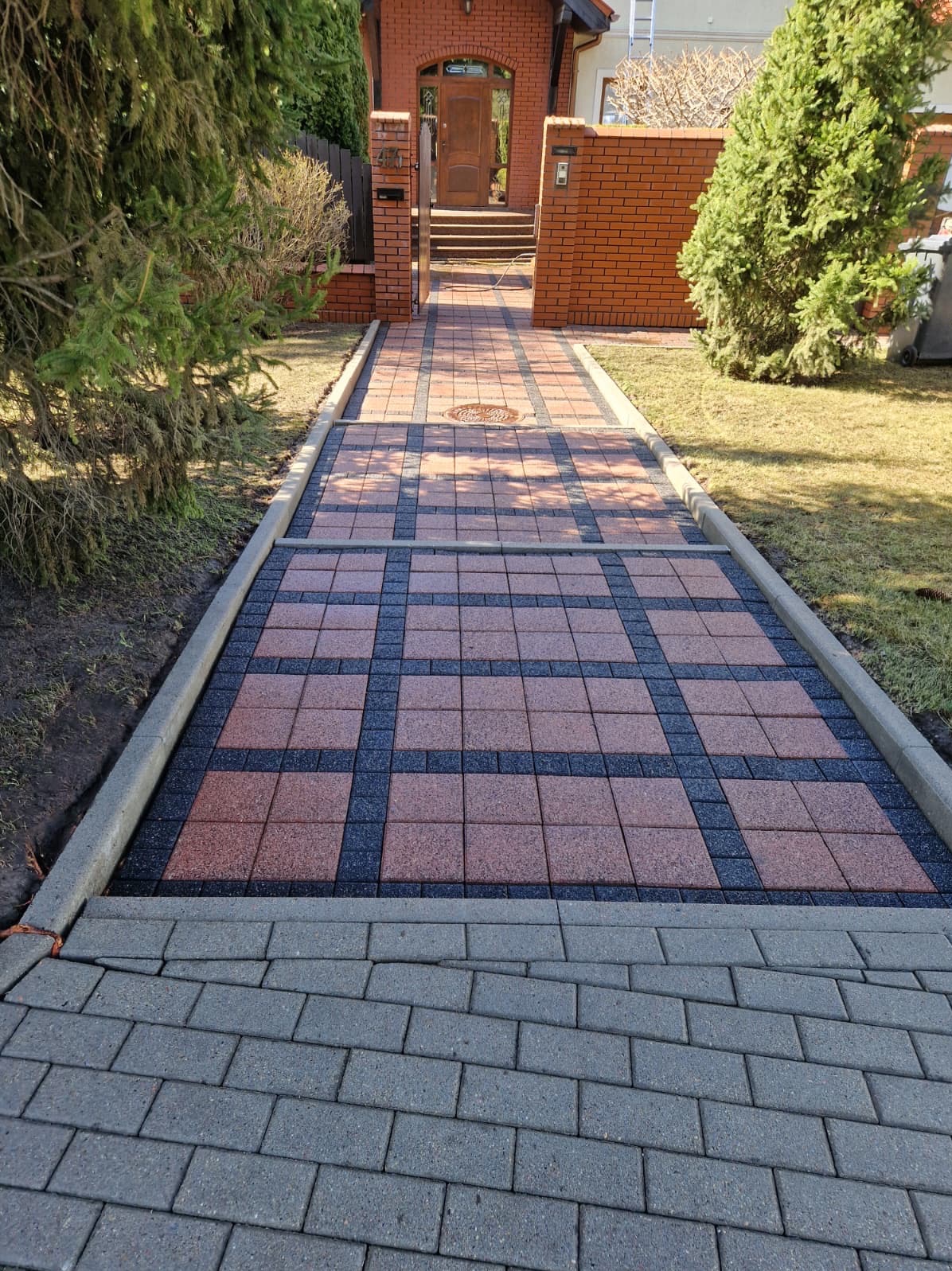 Ułożona kostka brukowa w geometryczny wzór prowadząca do domu z cegły, otoczona zielenią. Widok z perspektywy chodnika, w słoneczny dzień.