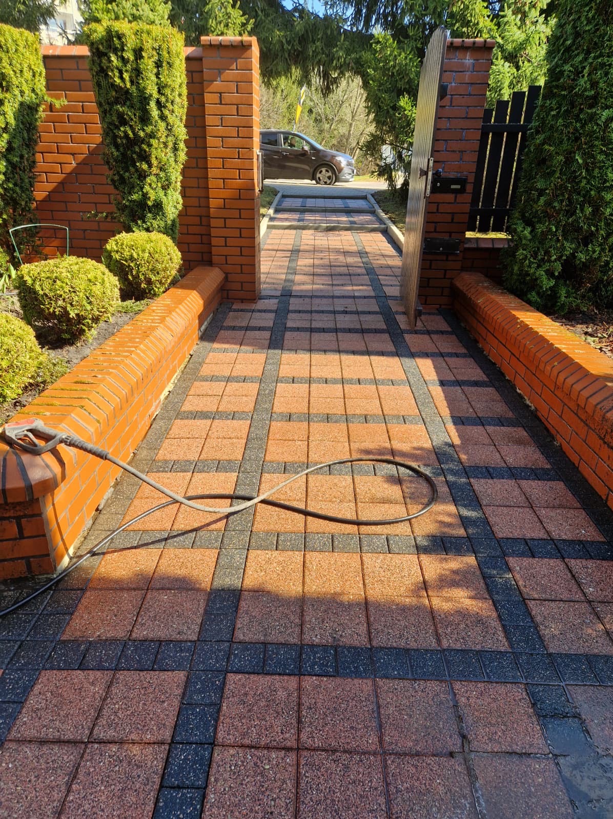 Ceglana ścieżka z kostki brukowej po czyszczeniu, widoczny wąż ciśnieniowy i mokra powierzchnia. Brama wejściowa i zieleń w tle.