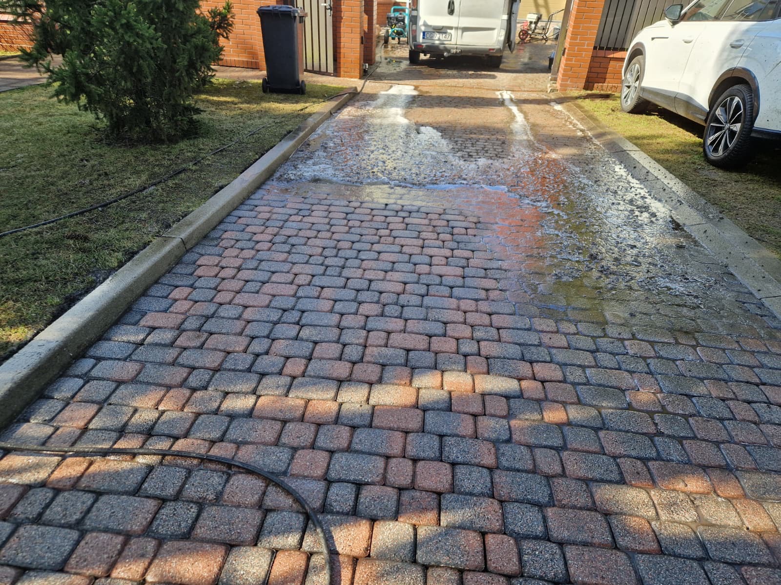 Częściowo umyta kostka brukowa na podjeździe, widoczny samochód i bus w tle. Woda spływa po czystej powierzchni, kontrastując z suchą, zabrudzoną częścią.