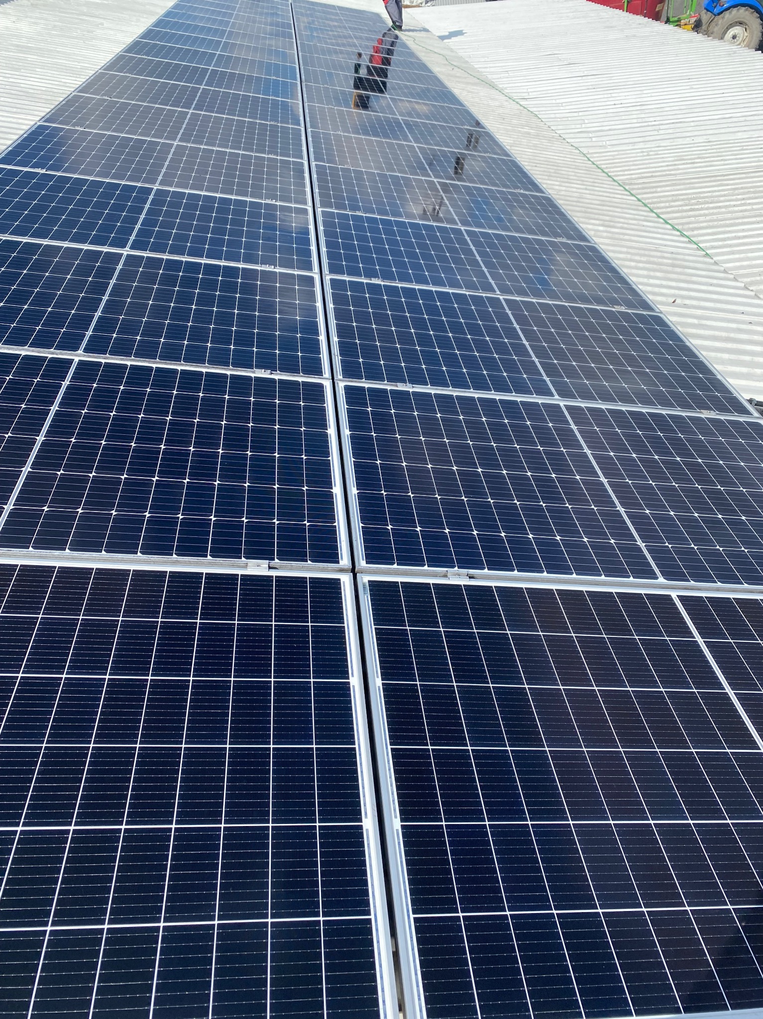 Błękitne panele słoneczne na falistym dachu, odbicie osoby w panelach, fragment traktora w tle, ujęcie z góry.