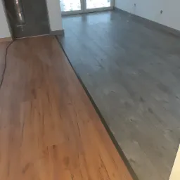 Fragment podłogi w trakcie układania: część wykończona panelami w odcieniach szarości, obok surowa podłoga przygotowana pod panele w kolorze drewna, widoczne niedokończone gniazdka elektryczne...