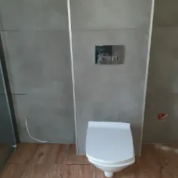 Nowoczesna toaleta wisząca z szarą, betonową okładziną ścienną i drewnopodobną podłogą, widoczna instalacja elektryczna w ścianie.