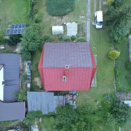 Zdjęcie z lotu ptaka przedstawiające dom z nowo pomalowaną czerwoną blachodachówką, otoczony zielonym trawnikiem i innymi budynkami, na jednym z budynków panele słoneczne.