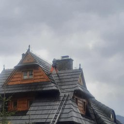 AlpinMal - Malowanie Dachów - Pracownik w pomarańczowych spodniach na dachu drewnianego domu w stylu góralskim z drabiną, prawdopodobnie wykonuje prace konserwatorskie lub czyszczenie, pochmurne niebo w tle.