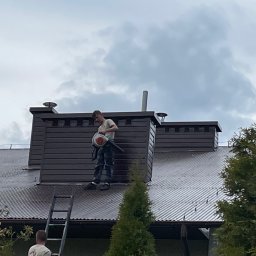 AlpinMal - Malowanie Dachów - Pracownik z dmuchawą na dachu domu podczas czyszczenia, widoczna drabina oparta o rynnę i komin z ciemnej blachy.