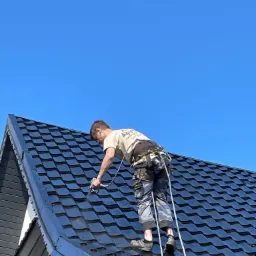 Pracownik w uprzęży zabezpieczającej, maluje natryskowo dach z blachodachówki w kolorze grafitowym, na tle błękitnego nieba.