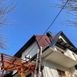 AlpinMal - Malowanie Dachów - Pracownik w kombinezonie czyści dach z brązowej dachówki za pomocą myjki ciśnieniowej, widok z dołu na dom z białymi ścianami, drewnianą pergolą i drabiną opartą o rynnę.