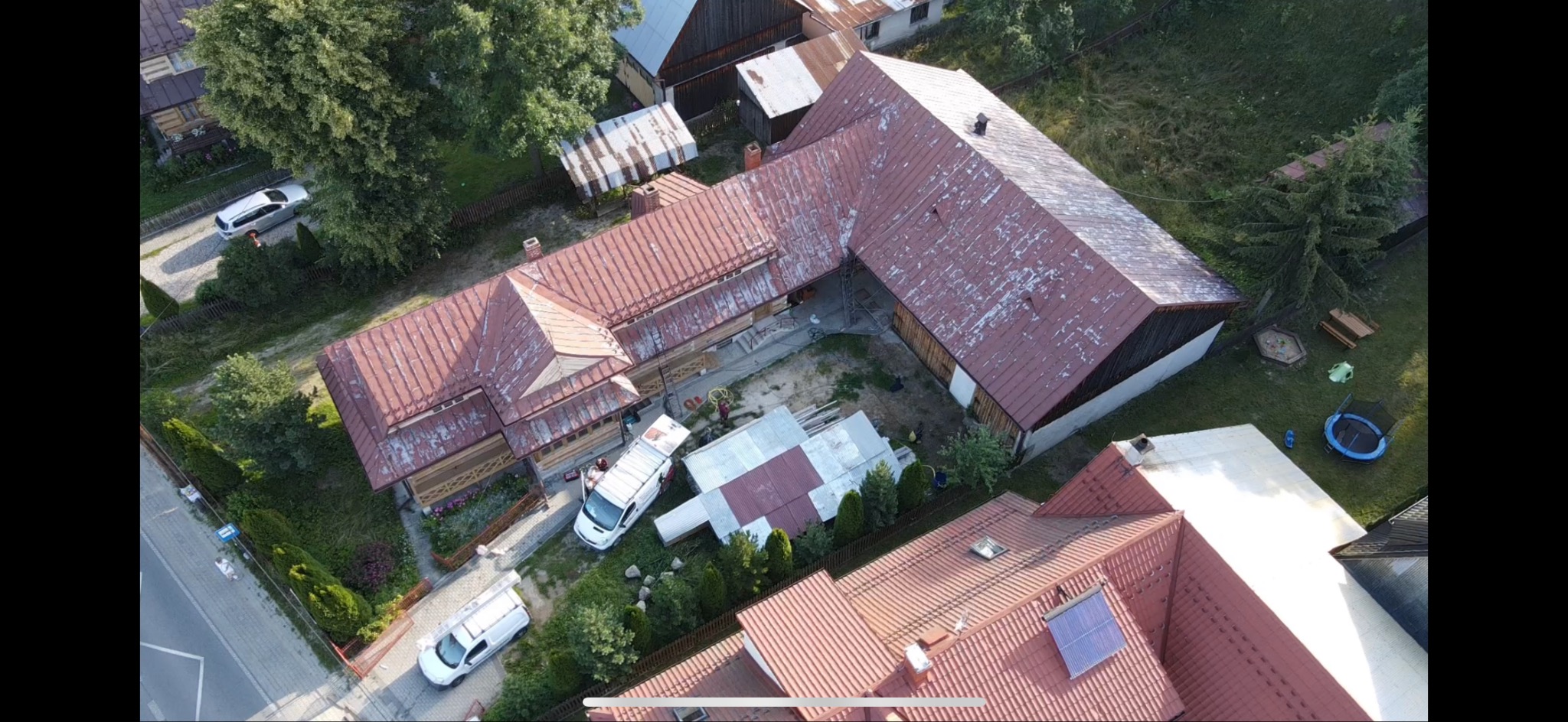 Widok z drona na duży, drewniany dom z czerwoną, miejscami wyblakłą blachodachówką. Dwa białe busy zaparkowane obok, jeden z otwartymi drzwiami. W tle inne budynki, drzewa i trawnik.