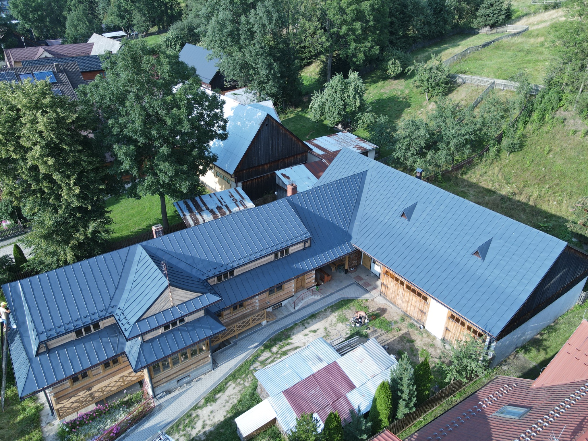 Panoramiczny widok z lotu ptaka na duży, drewniany dom z nowym, błyszczącym, szarym dachem blaszanym, otoczony zielenią drzew i trawników, z widocznymi ścieżkami z kostki brukowej i elementami...