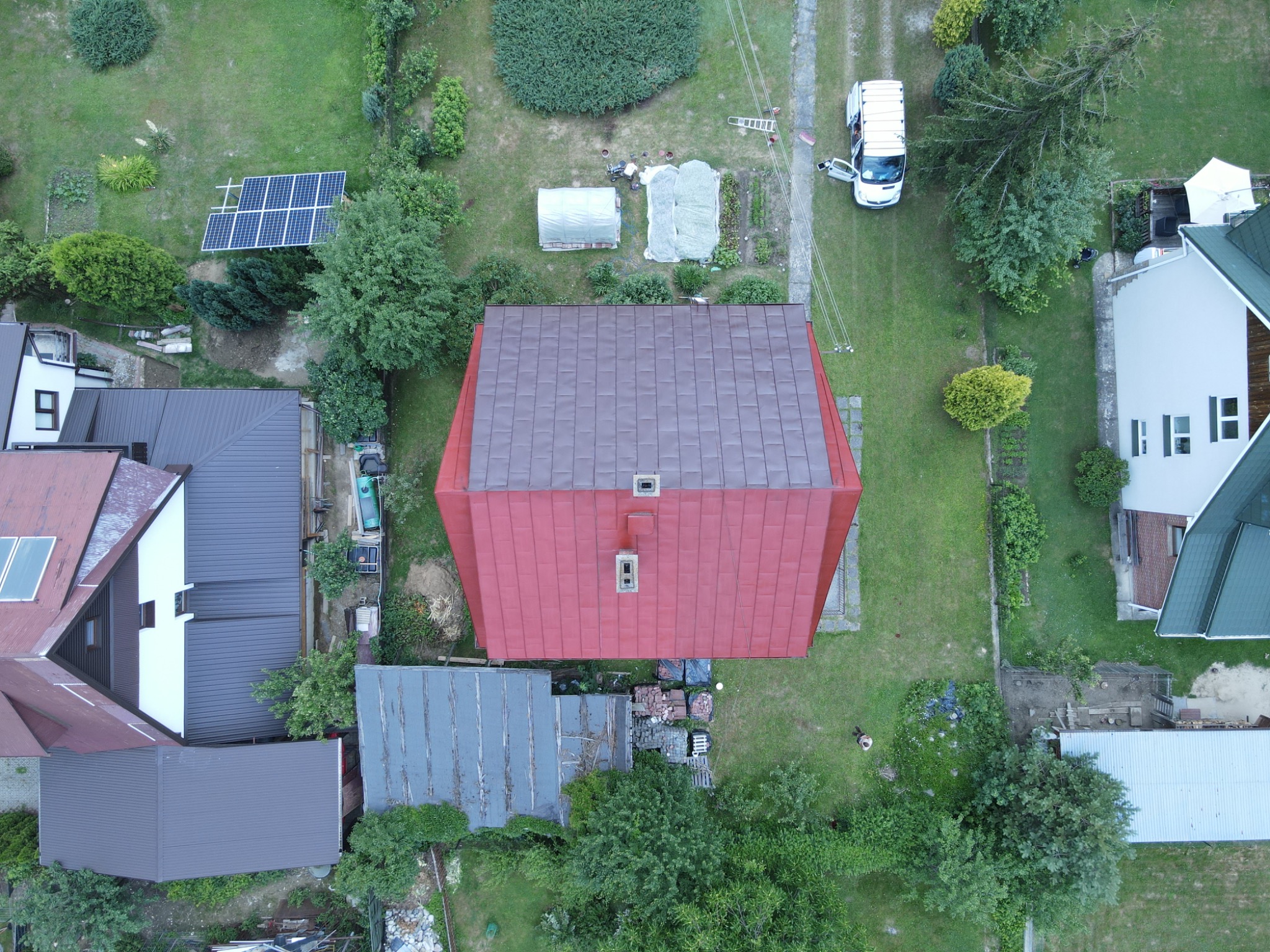 Zdjęcie z lotu ptaka przedstawiające dom z nowo pomalowaną czerwoną blachodachówką, otoczony zielonym trawnikiem i innymi budynkami, na jednym z budynków panele słoneczne.