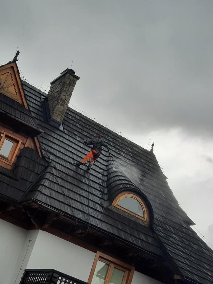 Pracownik w kombinezonie czyszczący ciśnieniowo ciemny, drewniany dach domu w stylu zakopiańskim z widocznym oknem mansardowym i kominem.