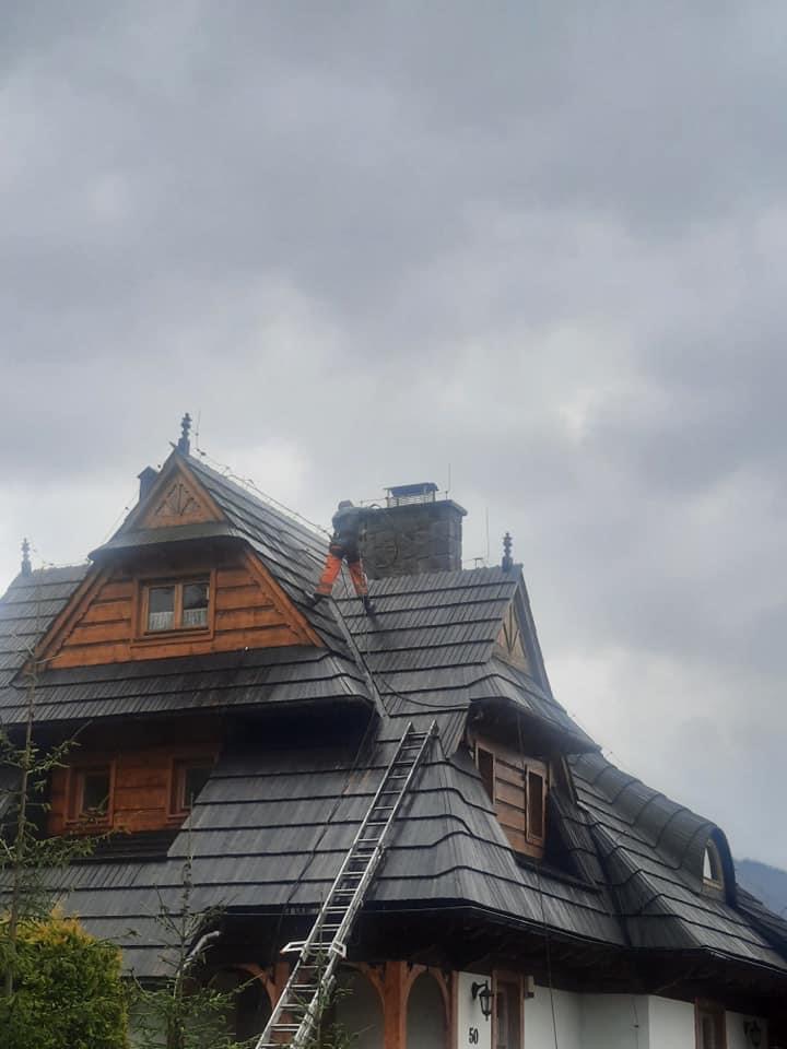 Pracownik w pomarańczowych spodniach na dachu drewnianego domu w stylu góralskim z drabiną, prawdopodobnie wykonuje prace konserwatorskie lub czyszczenie, pochmurne niebo w tle.