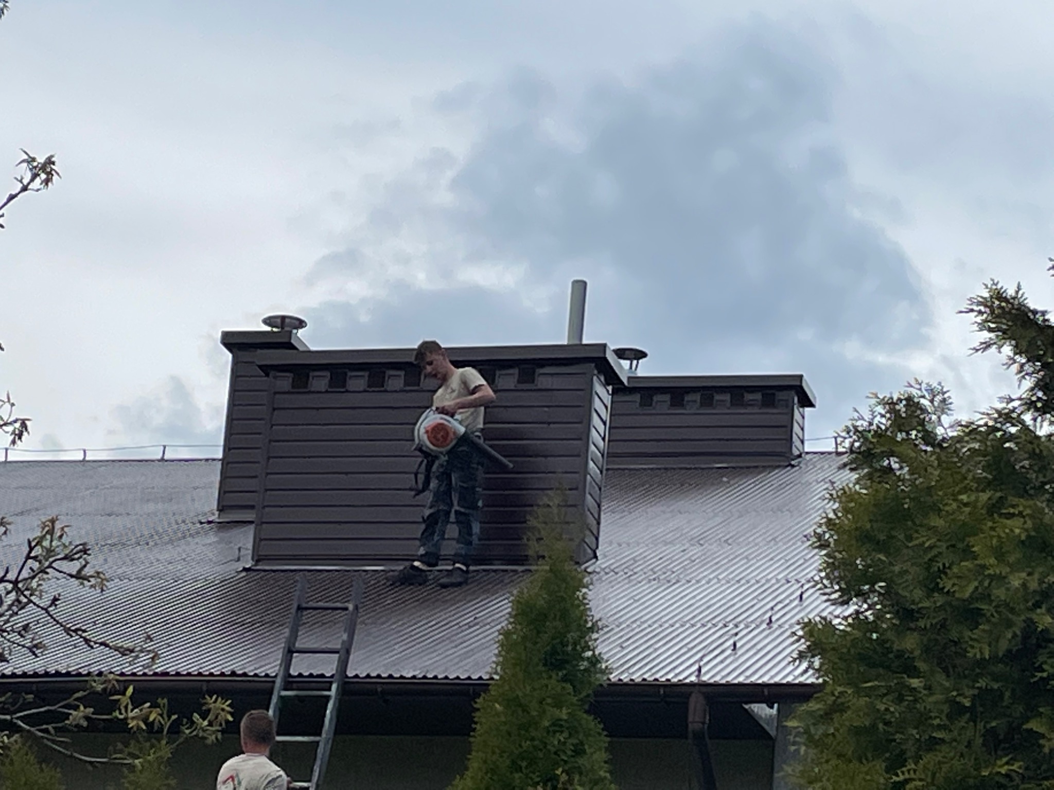 Pracownik z dmuchawą na dachu domu podczas czyszczenia, widoczna drabina oparta o rynnę i komin z ciemnej blachy.