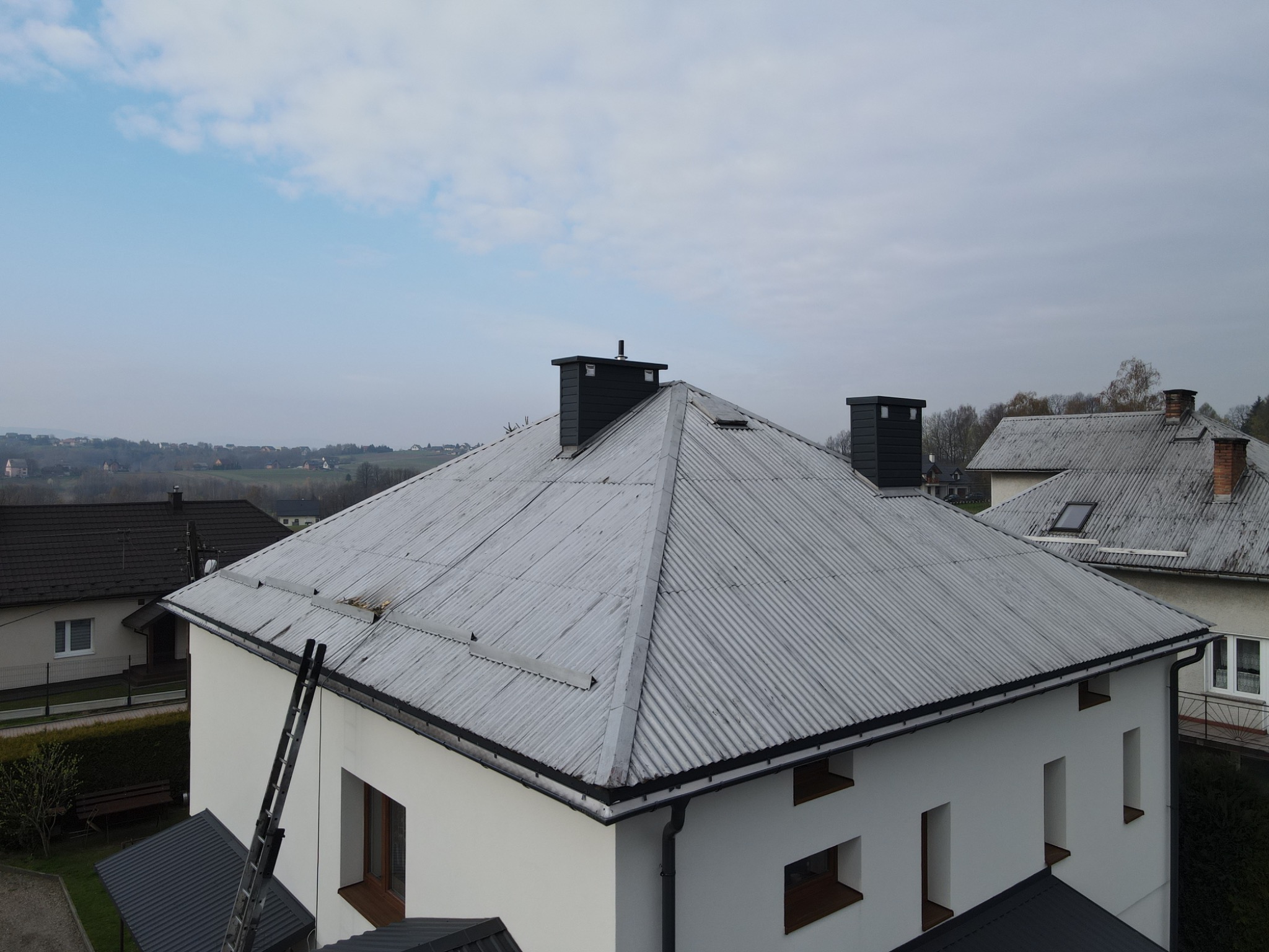 Widok z góry na biały dom z szarą, pofałdowaną blachą na dachu i czarnymi kominami, drabina oparta o rynnę, w tle zabudowa wiejska i zalesione wzgórza.