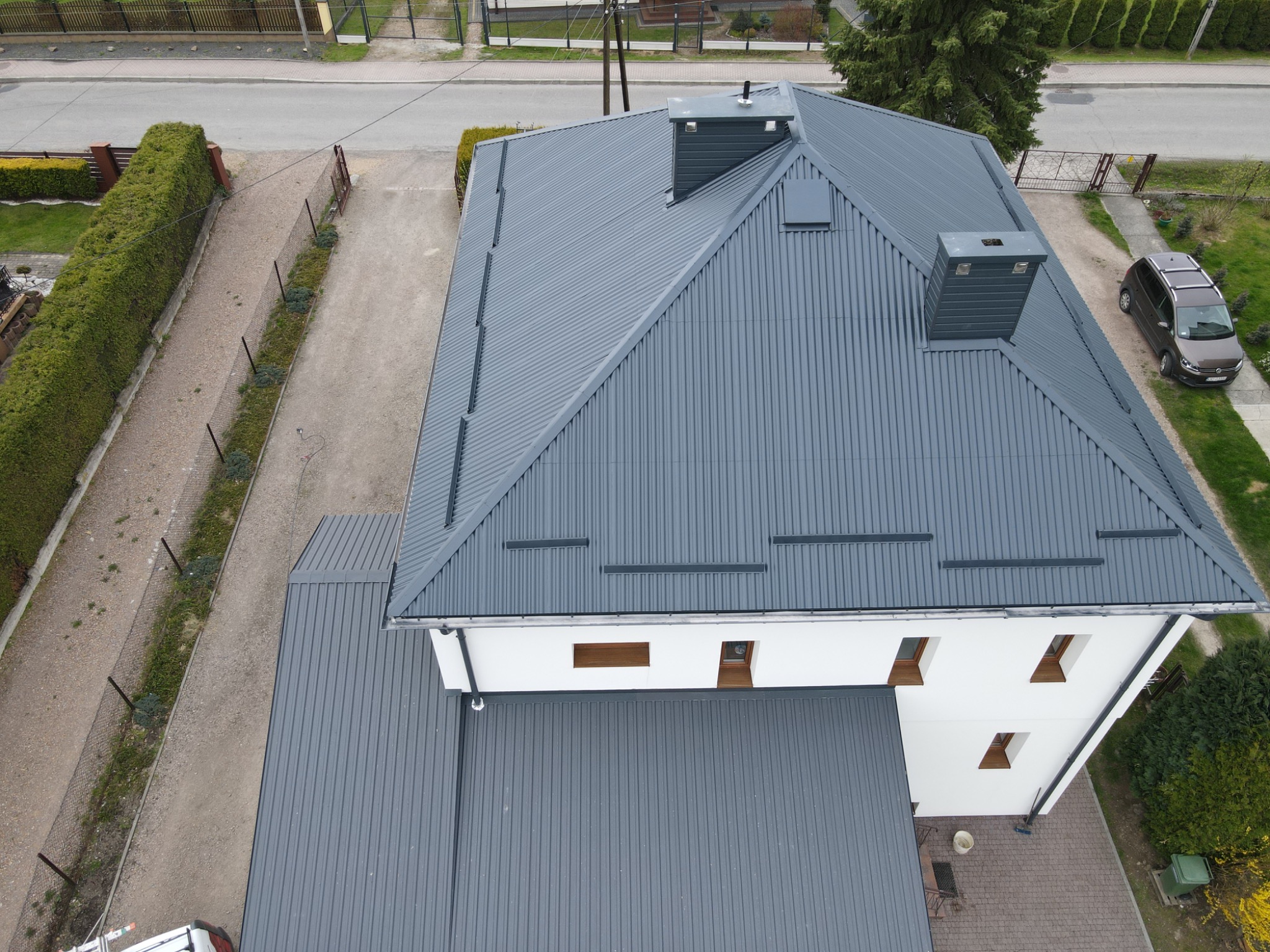 Widok z góry na dom z nowym, szarym dachem z blachy trapezowej i dwoma kominami, białą elewacją oraz brązowymi oknami, samochód osobowy zaparkowany obok domu.