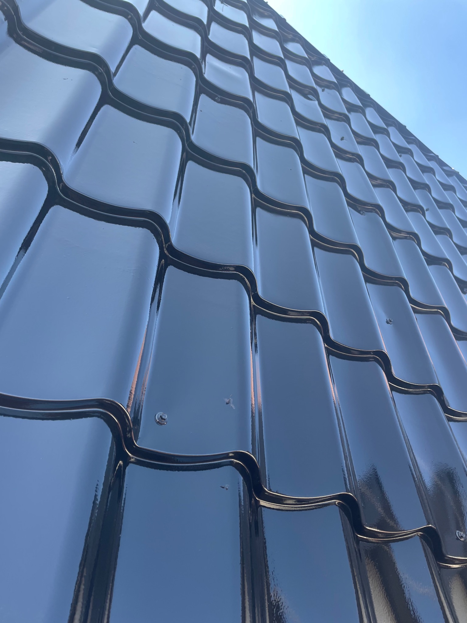 Błyszcząca, nowa, czarna dachówka ceramiczna ułożona na dachu widziana pod kątem z dołu, z widocznym fragmentem błękitnego nieba w tle.