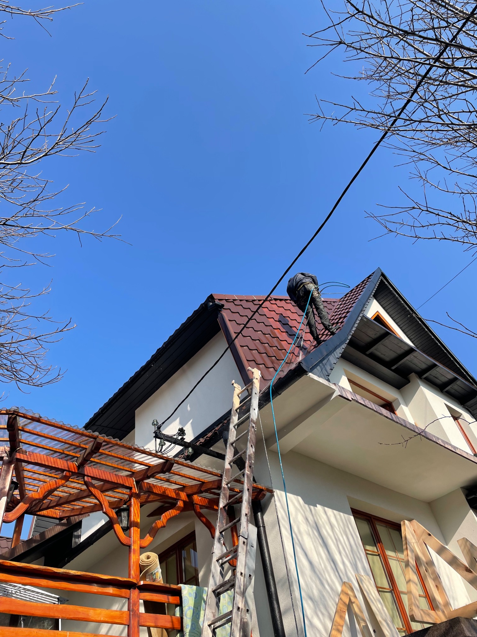Pracownik w kombinezonie czyści dach z brązowej dachówki za pomocą myjki ciśnieniowej, widok z dołu na dom z białymi ścianami, drewnianą pergolą i drabiną opartą o rynnę.