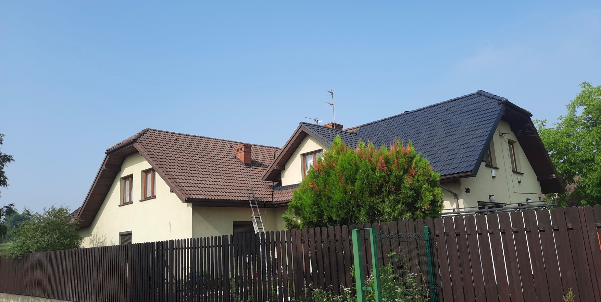 Dwa domy jednorodzinne za ciemnobrązowym płotem; jeden z brązowym dachem i beżową elewacją, drugi z ciemnoszarym dachem i kremową elewacją; na pierwszym domu oparta drabina