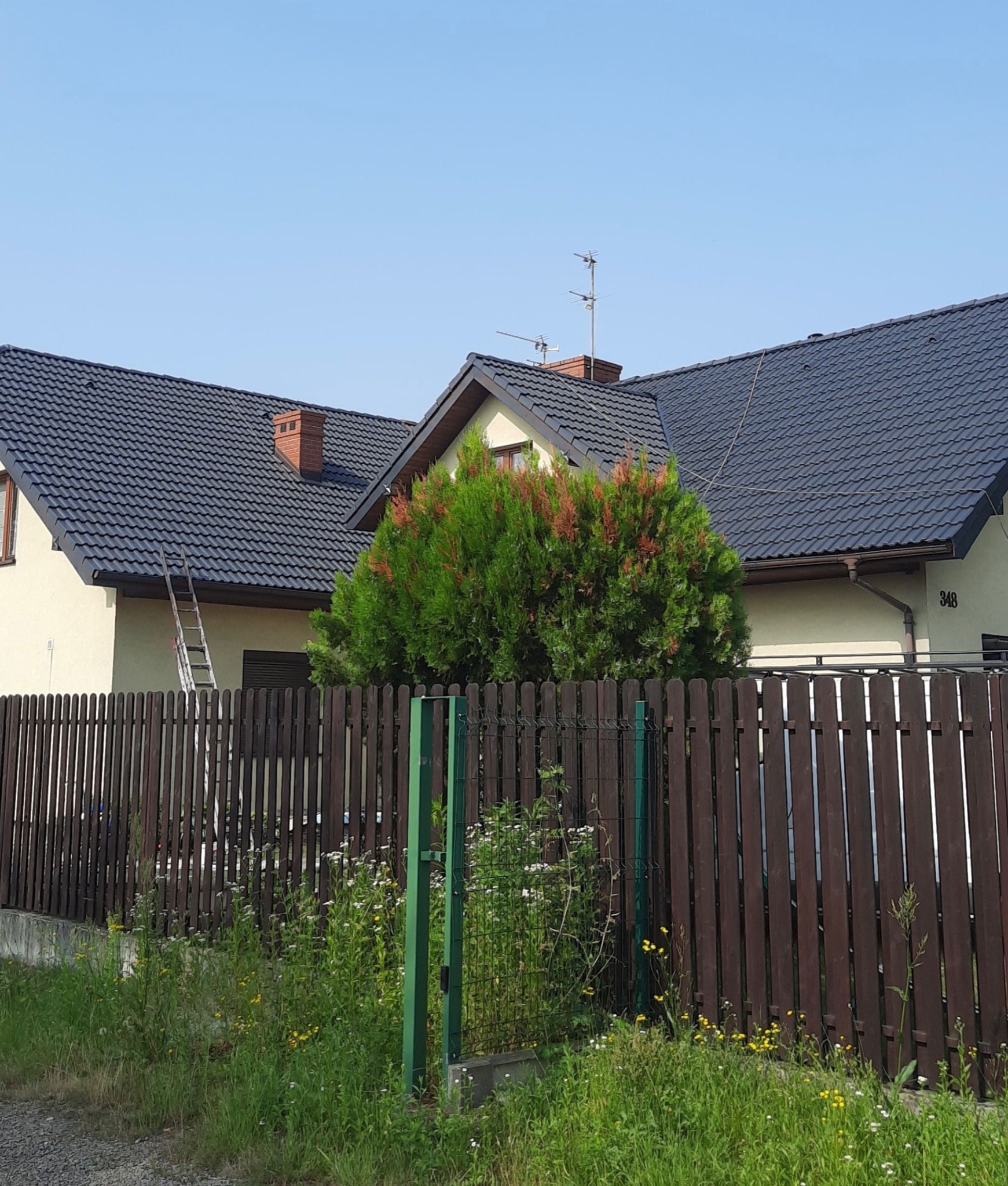 Dom jednorodzinny z ciemną dachówką, widoczna drabina oparta o rynnę na tle drewnianego płotu i zieleni.