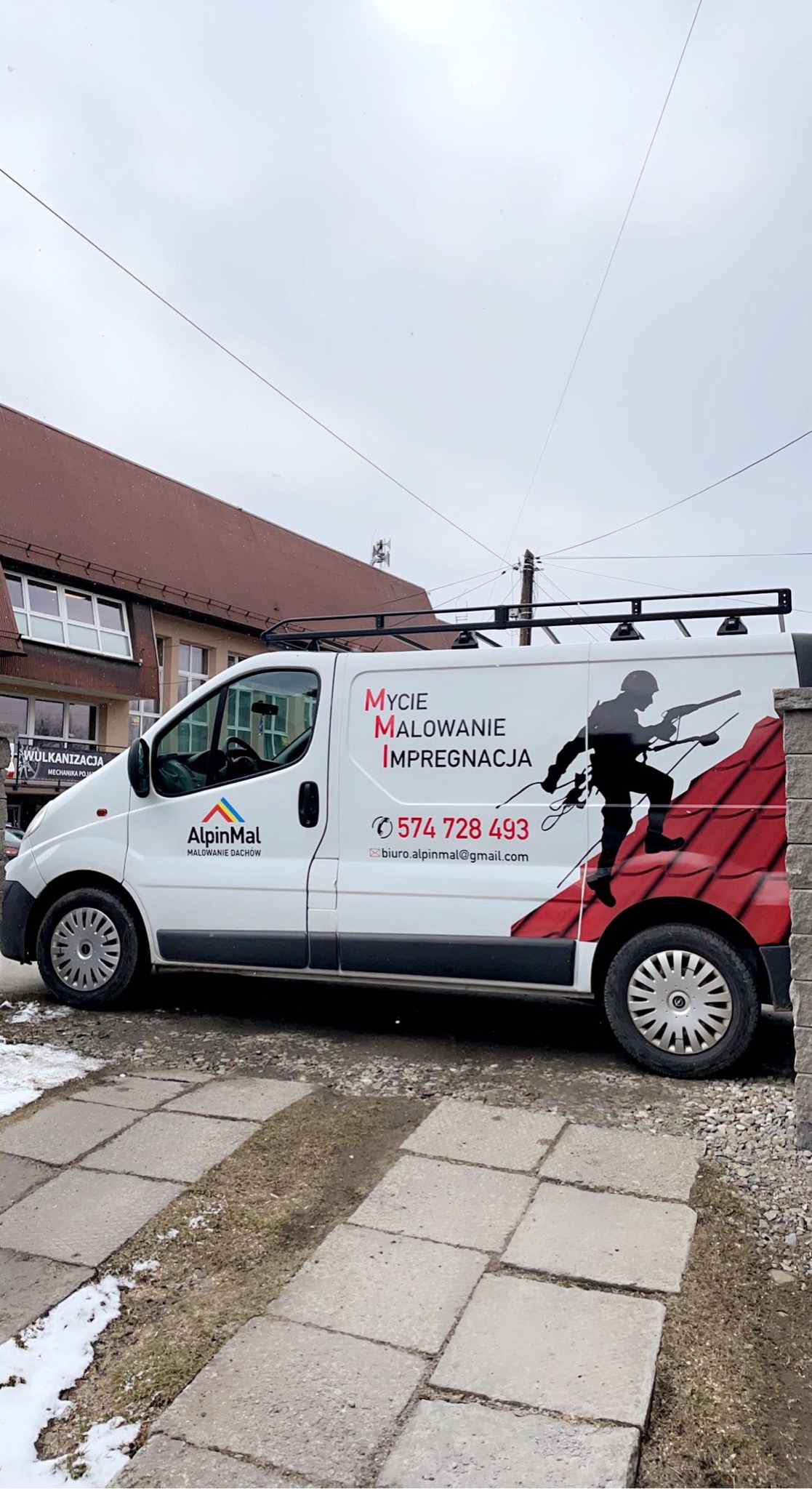 Biały samochód dostawczy z logo firmy AlpinMal, oferującej mycie, malowanie i impregnację dachów, zaparkowany na chodniku z kostki brukowej przed budynkiem.
