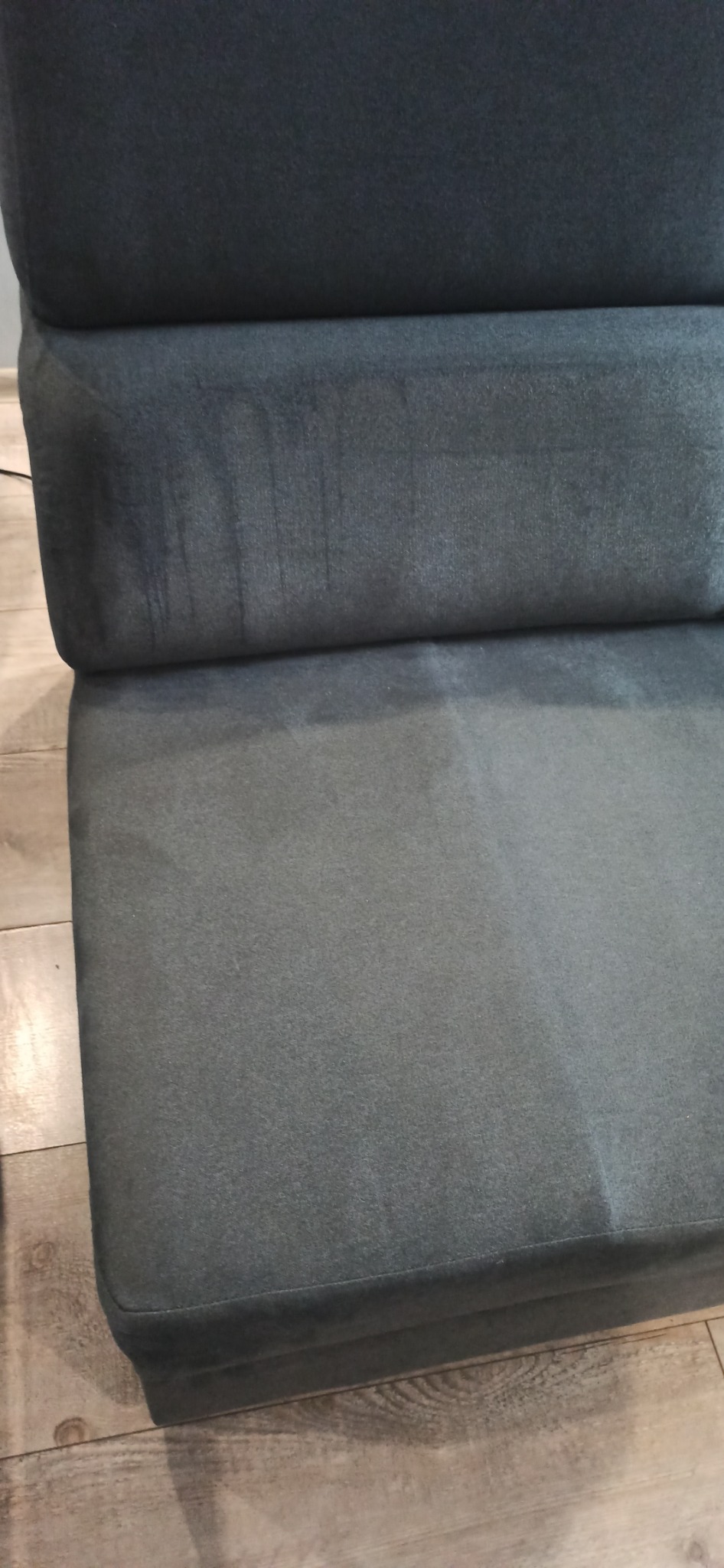 Fragment ciemnoszarej, welurowej kanapy z widocznymi, świeżymi plamami po czyszczeniu tapicerki, ukazujący różnicę w kolorze przed i po zabiegu.