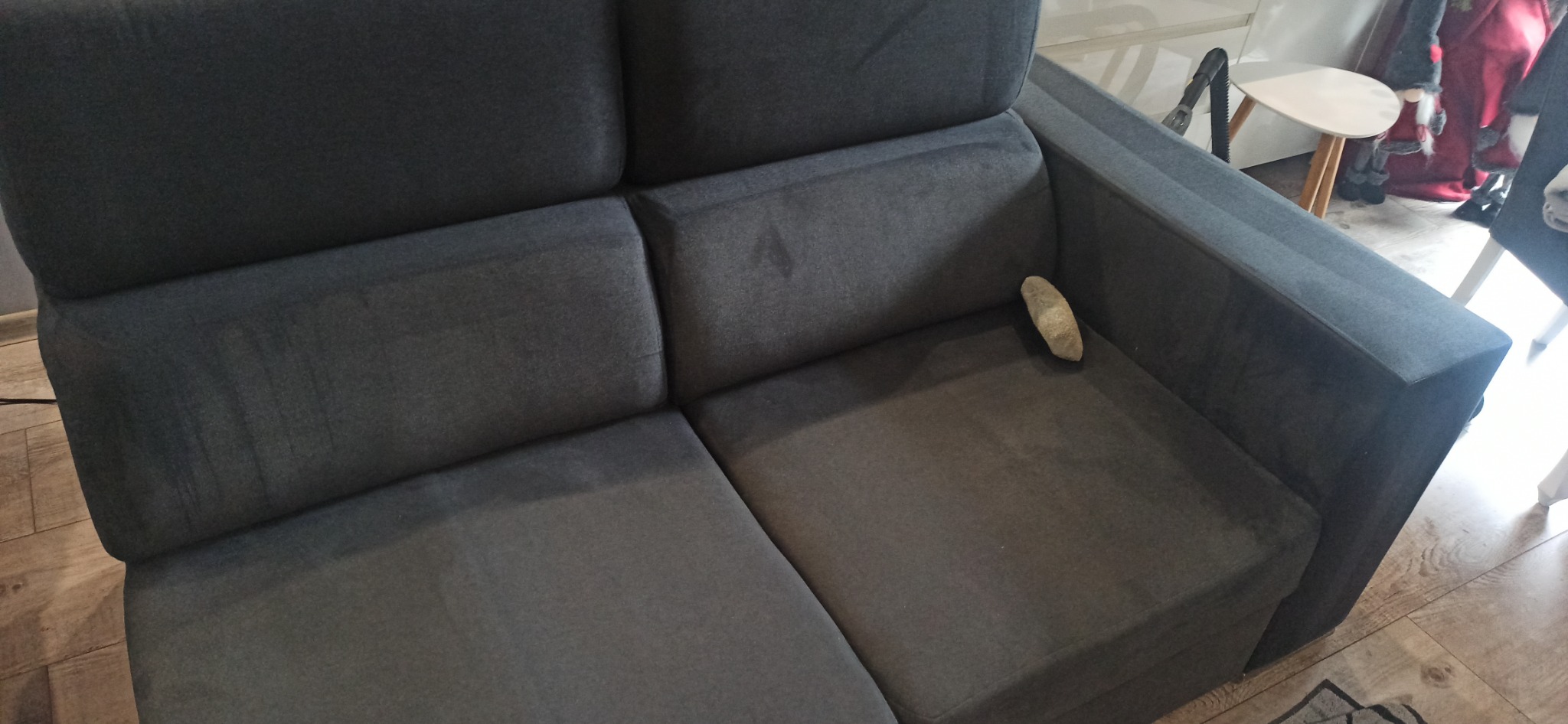 Ciemnoszara kanapa z plamą na siedzisku, na której leży mały kamień. Widoczny fragment podłogi i elementy wystroju w tle.