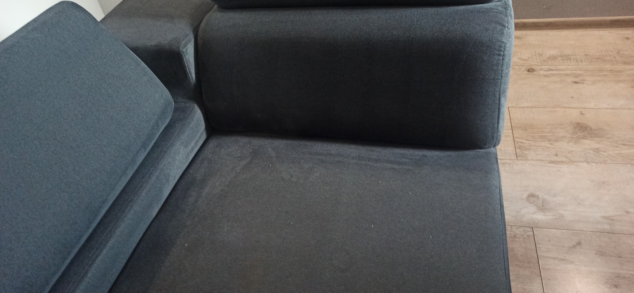 Fragment szarej, welurowej kanapy narożnej z widocznymi śladami użytkowania, na tle paneli podłogowych.