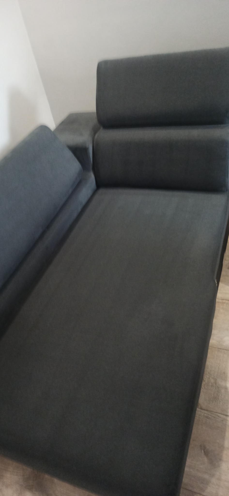 Szara, rozłożona kanapa z poduszką na boku, ustawiona na drewnianej podłodze w rogu pokoju.