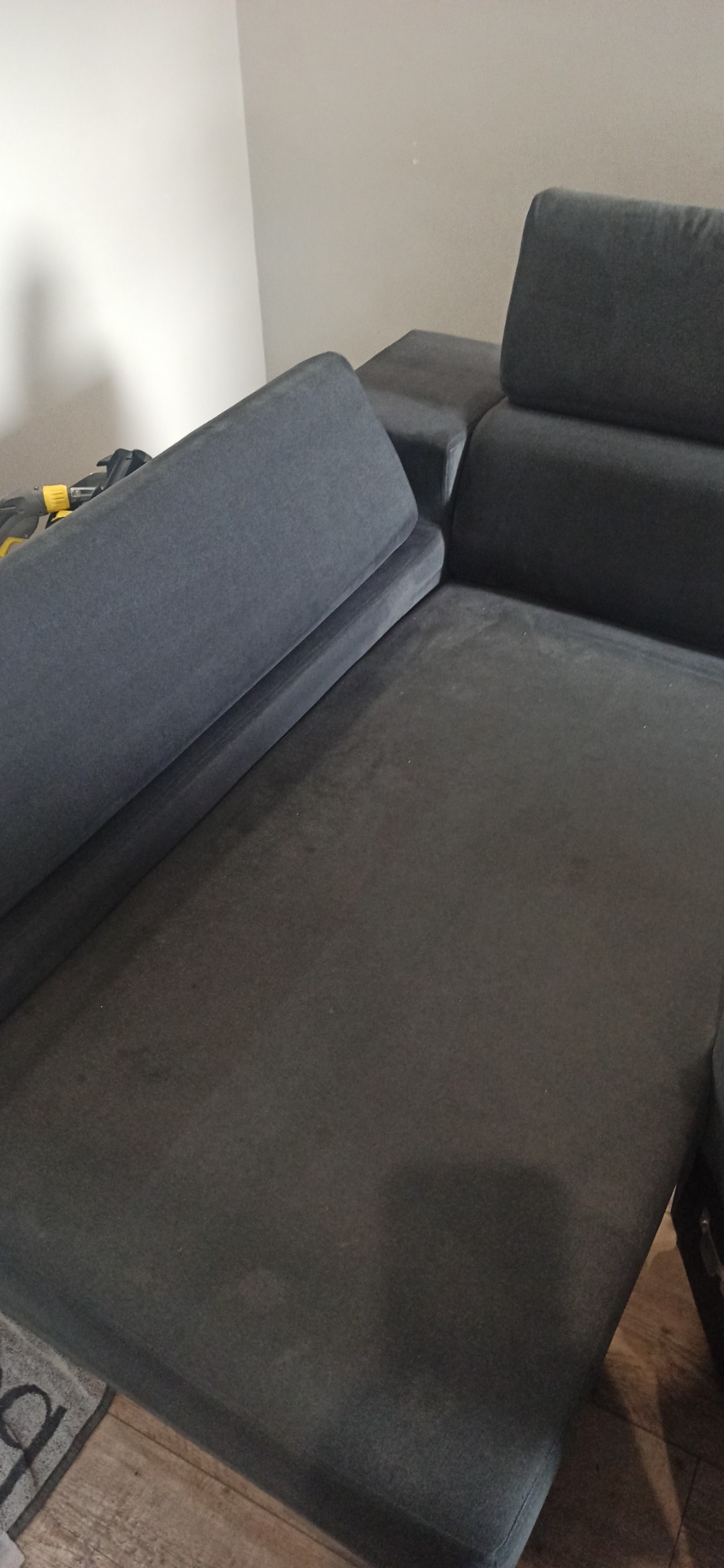 Ciemnoszara, modułowa sofa z podniesionym oparciem, widoczne drobne zabrudzenia na siedzisku, obok leży żółty odkurzacz piorący.
