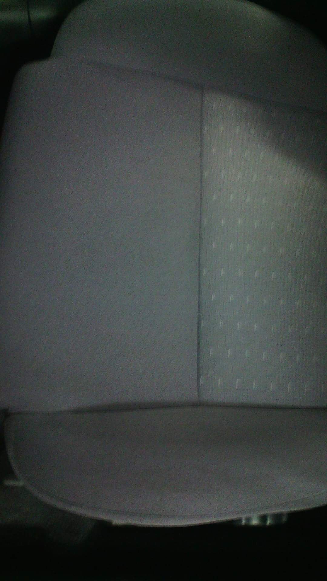 Szare, materiałowe obicie fotela samochodowego z widocznym szwem i wzorem w kropki po prawej stronie, uchwycone w słabym oświetleniu.