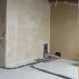 Pokój w trakcie prac wykończeniowych: ściany po szpachlowaniu, widoczne instalacje wodne i elektryczne w ścianie, rury na podłodze, surowy stan.