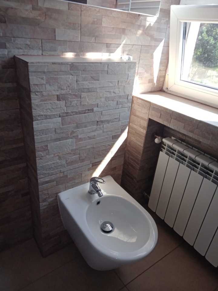Bidet w łazience wykończonej płytkami imitującymi kamień, z widocznym grzejnikiem i oknem z widokiem na zieleń.