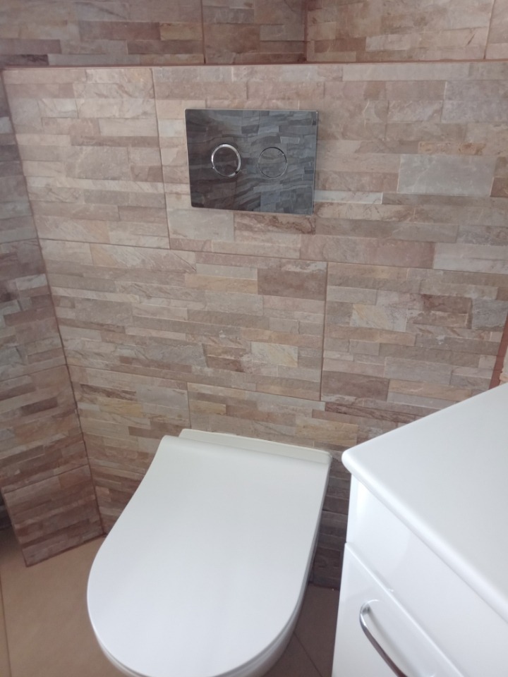 Wisząca toaleta z białym siedziskiem i chromowanym panelem spłukującym na ścianie wyłożonej płytkami imitującymi kamień.