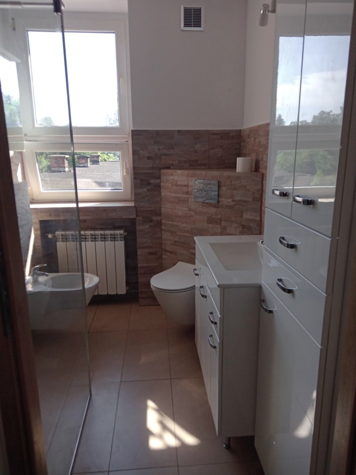 Jasna łazienka z brązowymi akcentami: beżowe płytki imitujące kamień na ścianach, białe meble łazienkowe z chromowanymi uchwytami, bidet, toaleta i grzejnik pod oknem.