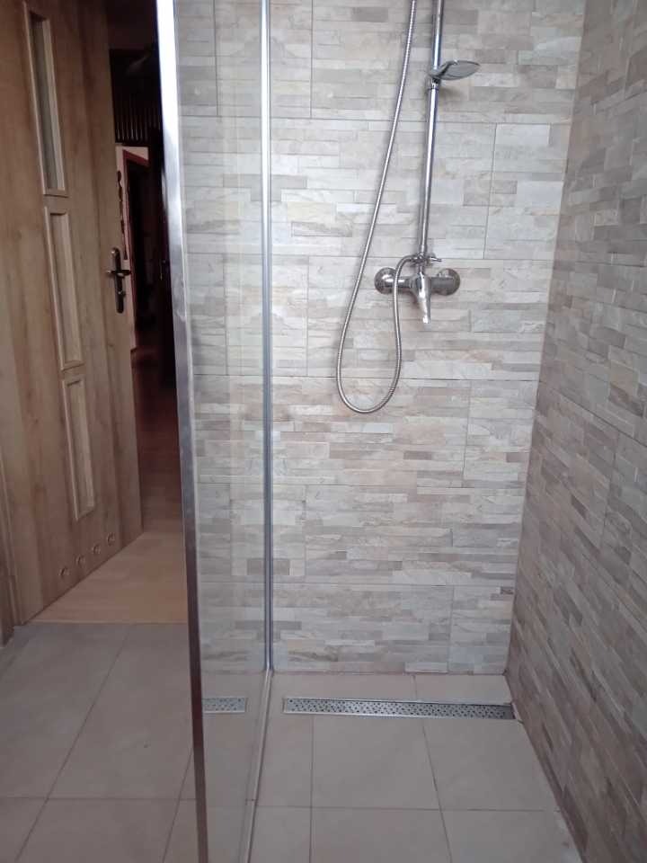 Nowoczesna kabina prysznicowa z odpływem liniowym, wykończona płytkami imitującymi kamień w odcieniach beżu i szarości, widoczna częściowo otwarta drewniana rama drzwi.