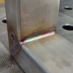 Spoina pachwinowa na profilu stalowym, z widocznymi kolorami hartowania, umieszczona na stole spawalniczym z rastrem otworów.