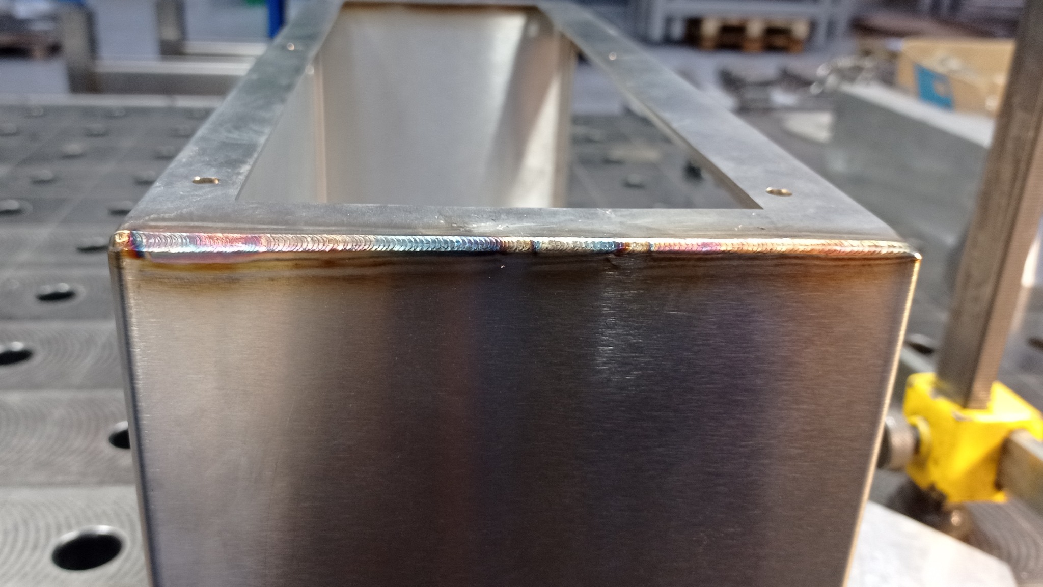 Metalowa konstrukcja skrzynkowa ze spoinami TIG o charakterystycznym zabarwieniu na krawędziach, widoczne ślady obróbki na powierzchni metalu, tło stanowią elementy warsztatu.