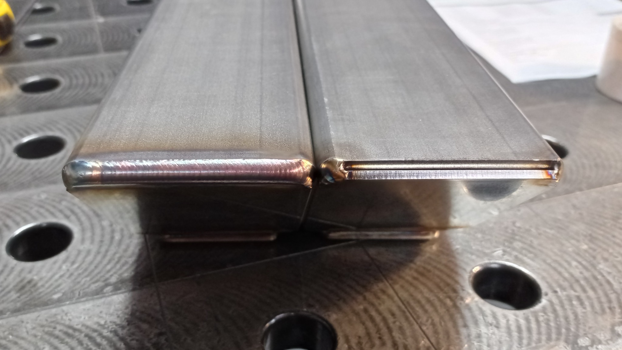 Zbliżenie na dwa spawane, prostokątne profile metalowe, leżące na perforowanym stole spawalniczym. Widoczne spawy na krawędziach.