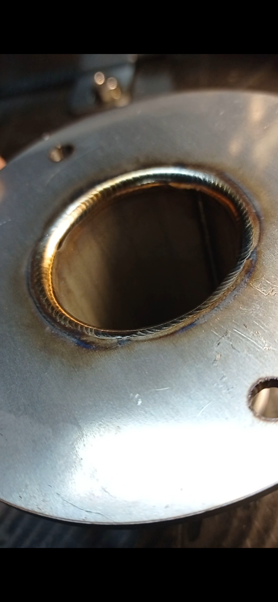 Szczegółowe zbliżenie na spawany element z metalu, widoczny precyzyjny szew spawalniczy z charakterystycznym złotym odcieniem, otwór w elemencie prowadzi w głąb konstrukcji.