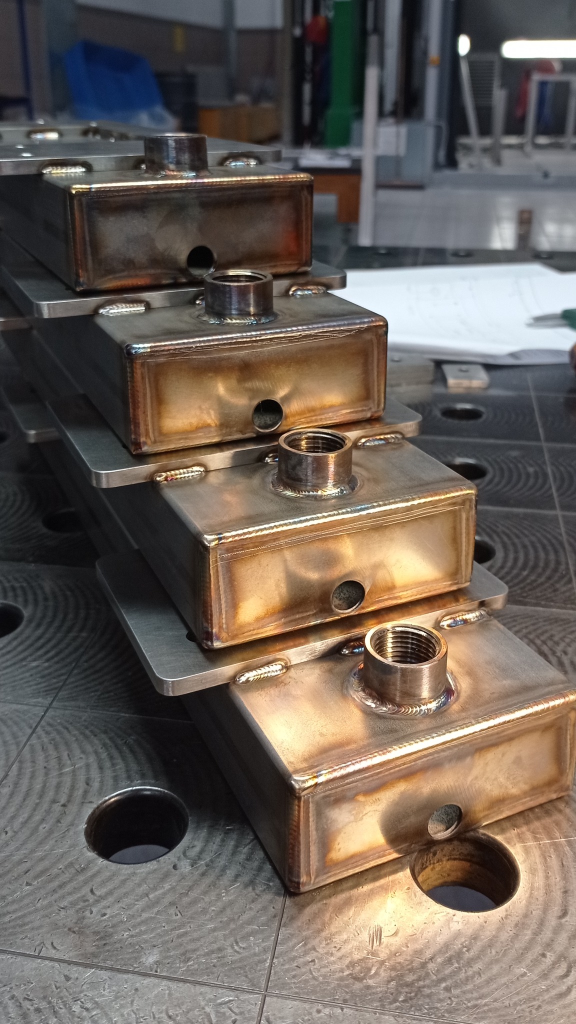 Cztery metalowe, spawane zbiorniki ułożone jeden na drugim, z widocznymi śladami spawania i otworami gwintowanymi, leżące na perforowanym stole spawalniczym.