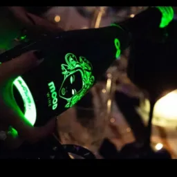 Osoba nalewa wino z czarnej butelki z zielonym, świecącym logo i napisem 'mood' do kieliszka podczas imprezy.