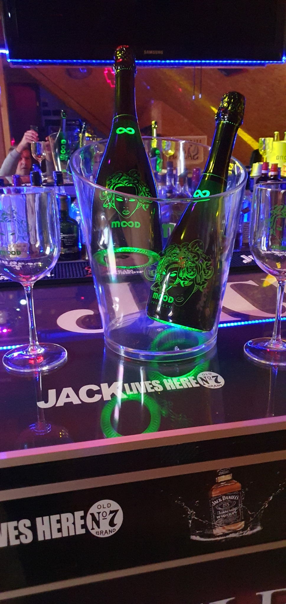 Dwie butelki prosecco marki Mood w przezroczystym wiaderku z lodem, obok dwa puste kieliszki do wina, na barze z neonowym oświetleniem i napisem Jack Lives Here.