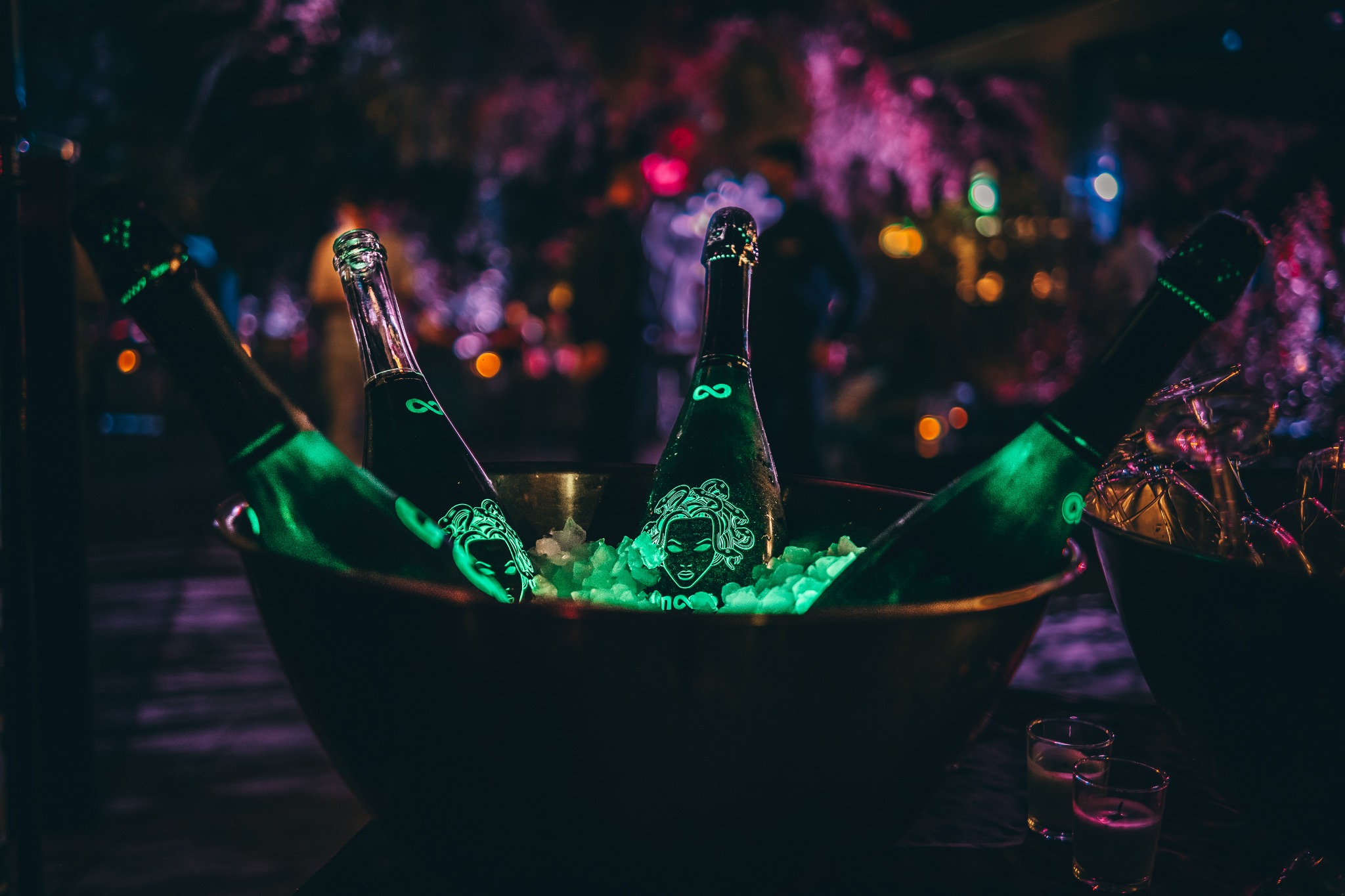 Cztery butelki szampana z zielonym podświetleniem w metalowej misie wypełnionej lodem, na butelkach widoczny motyw głowy Meduzy, w tle rozmyte światła imprezowe.