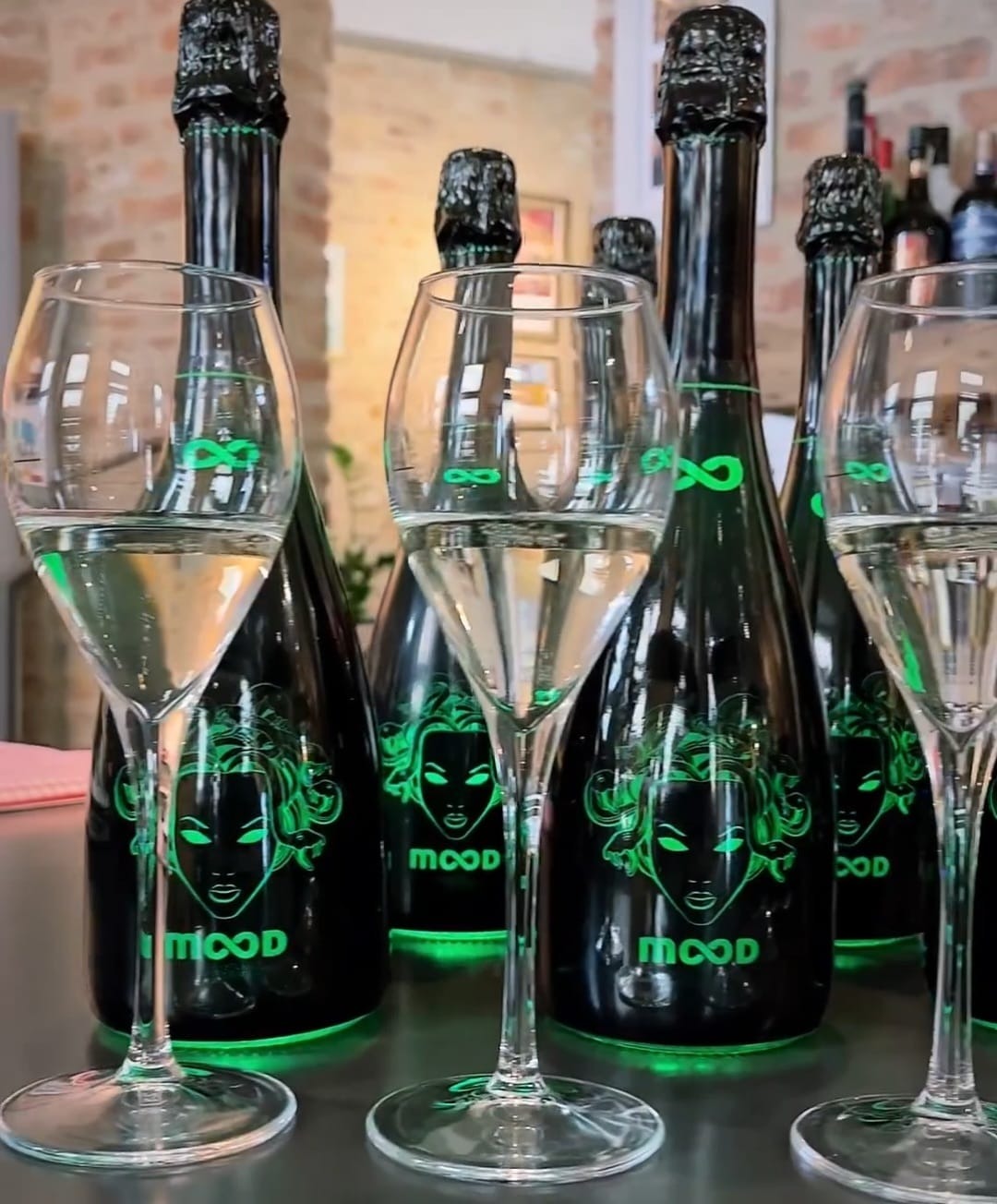 Ustawione na blacie czarne butelki szampana z zielonym neonowym nadrukiem twarzy kobiety i napisem 'MOOD', obok stoją kieliszki wypełnione płynem.
