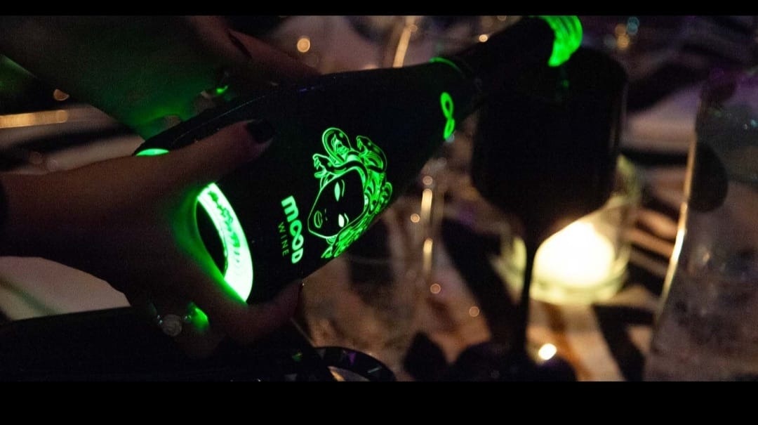 Osoba nalewa wino z czarnej butelki z zielonym, świecącym logo i napisem 'mood' do kieliszka podczas imprezy.